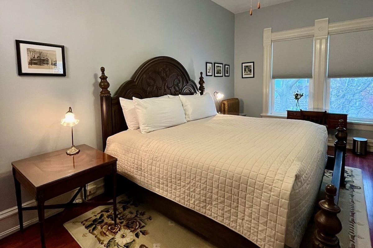 A bedroom with a bed, a nightstand, a floor lamp, a ceiling fan, and a window with a view.