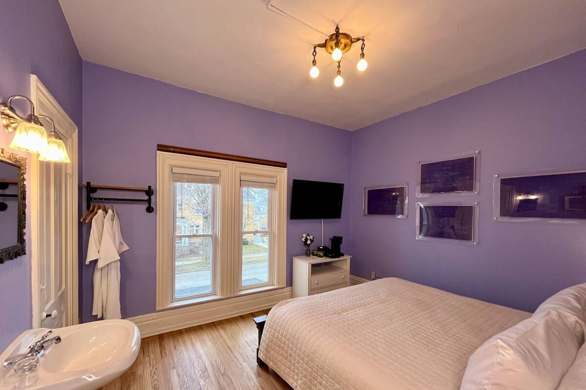A bedroom with a bed, a bedside table, a TV, a window, and a bathroom with a sink and mirror.