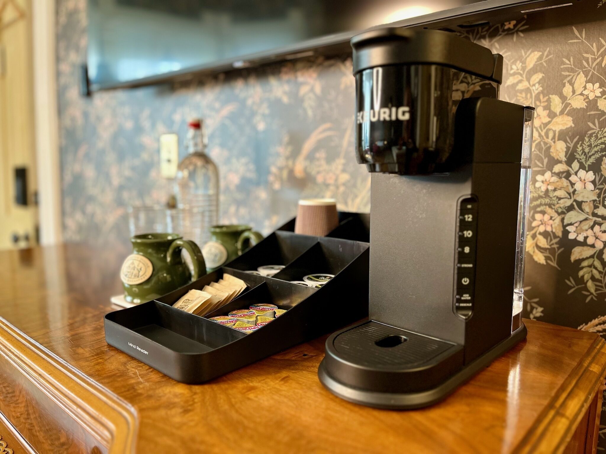 A coffee maker with a tray of tea bags and a green mug on a wooden countertop.