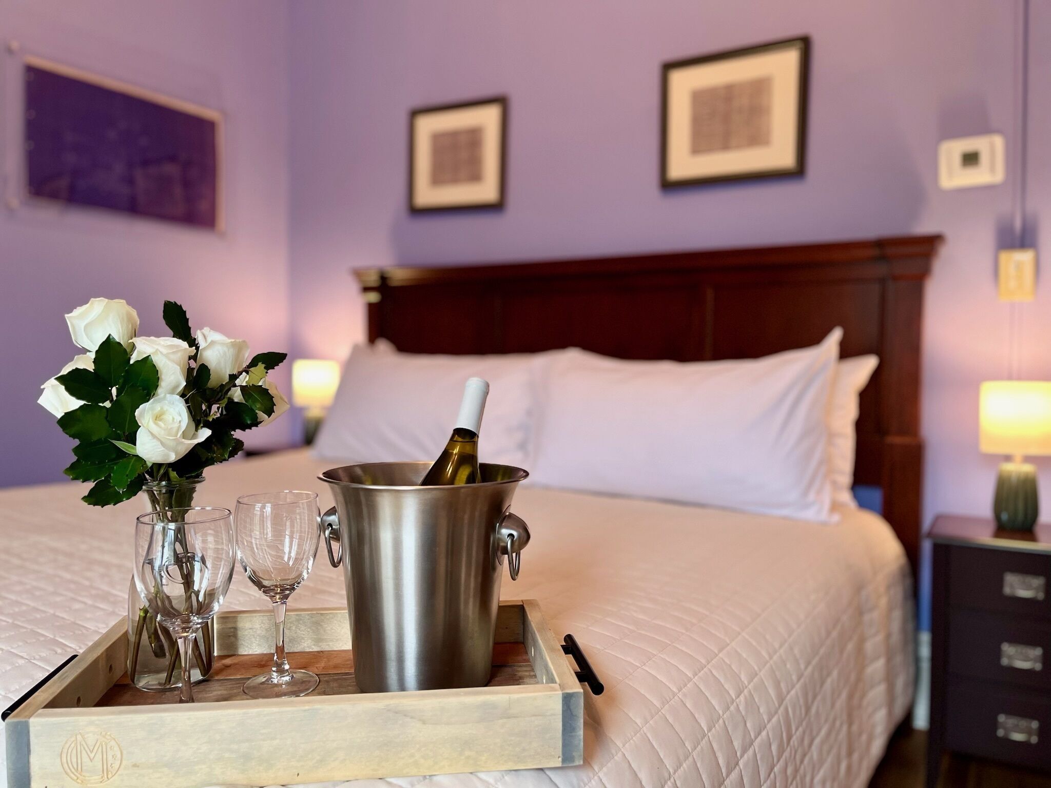 A hotel room with a bed, a tray with a bottle of champagne, two wine glasses, and a vase of white roses.