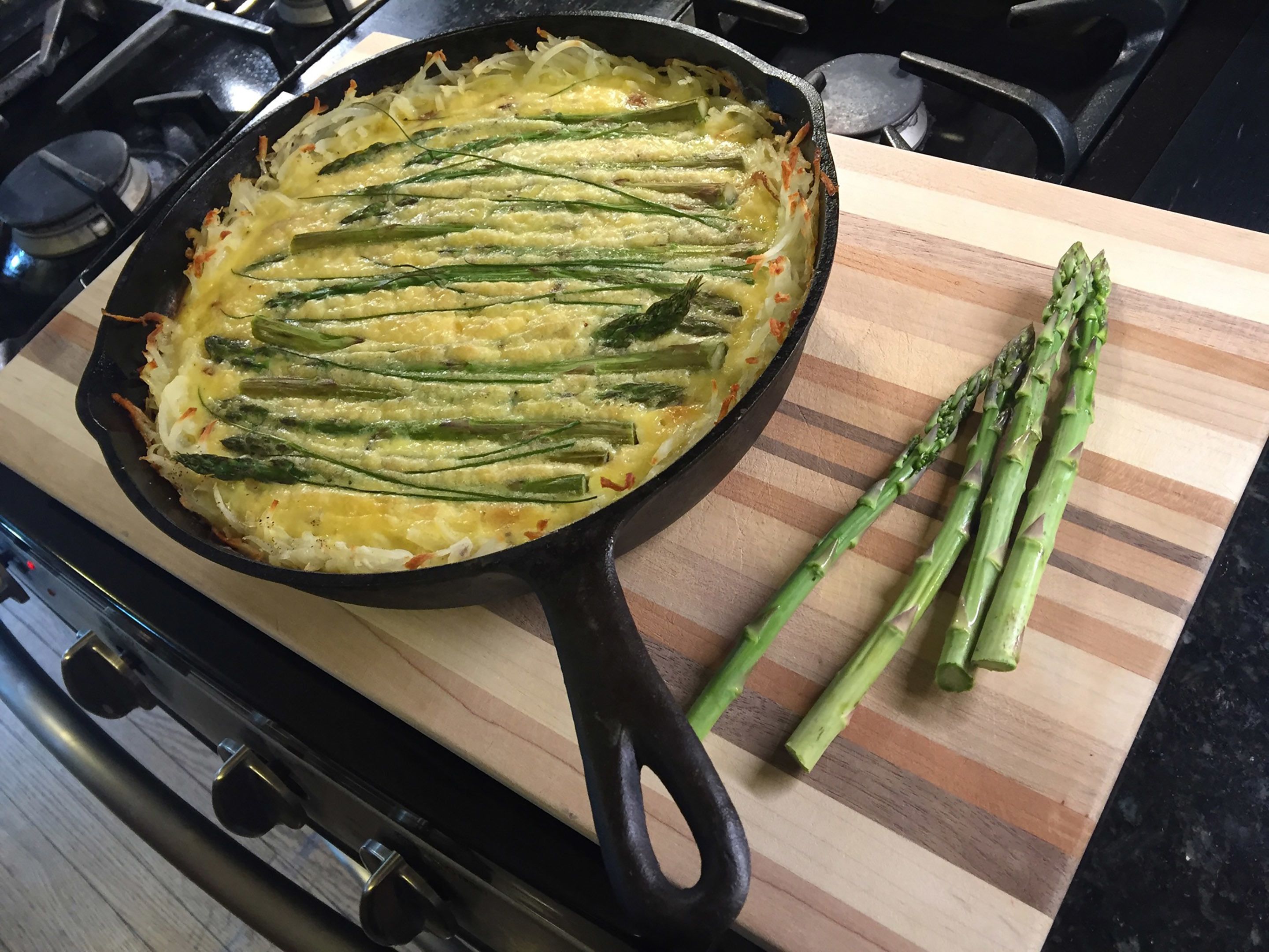 A cast iron skillet with a dish containing asparagus and potatoes on a wooden cutting board.