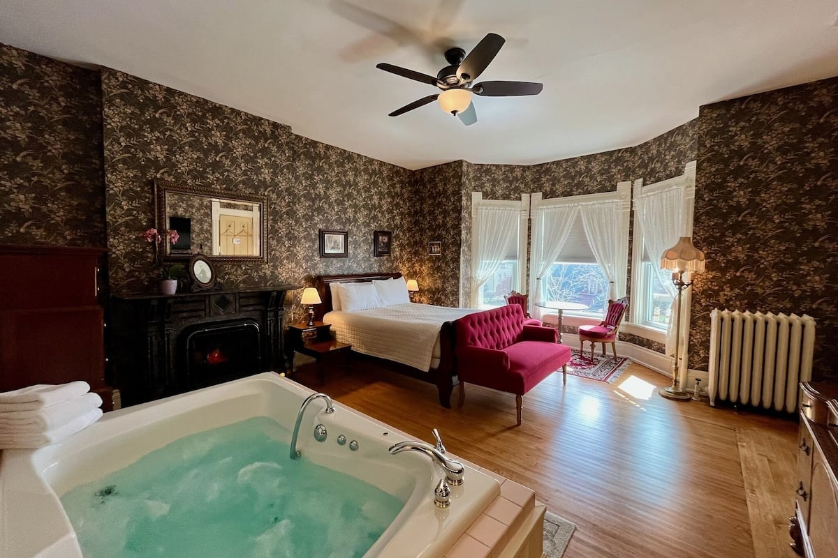 A hotel room with a large bathtub, a bed with a red chair, a fireplace, and a view of the outdoors through the windows.