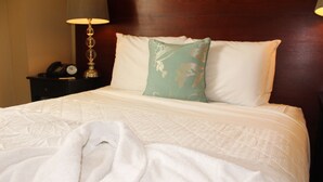 Una cama bien tendida con una colcha blanca y un almohadón verde, una lámpara de noche y una mesita de noche con un reloj.