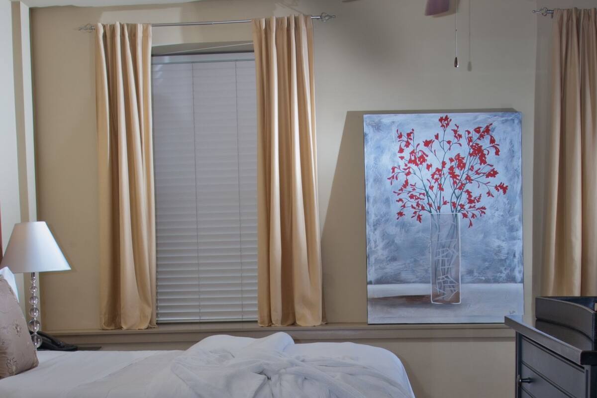 A bedroom with a bed, a dresser, a ceiling fan, a window with curtains, and a painting of a vase with flowers.