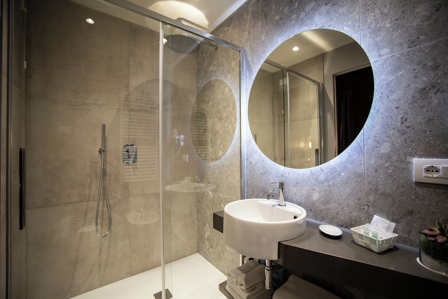 Un baño moderno con ducha envuelta en cristal, un espejo redondo y un lavamanos blanco.