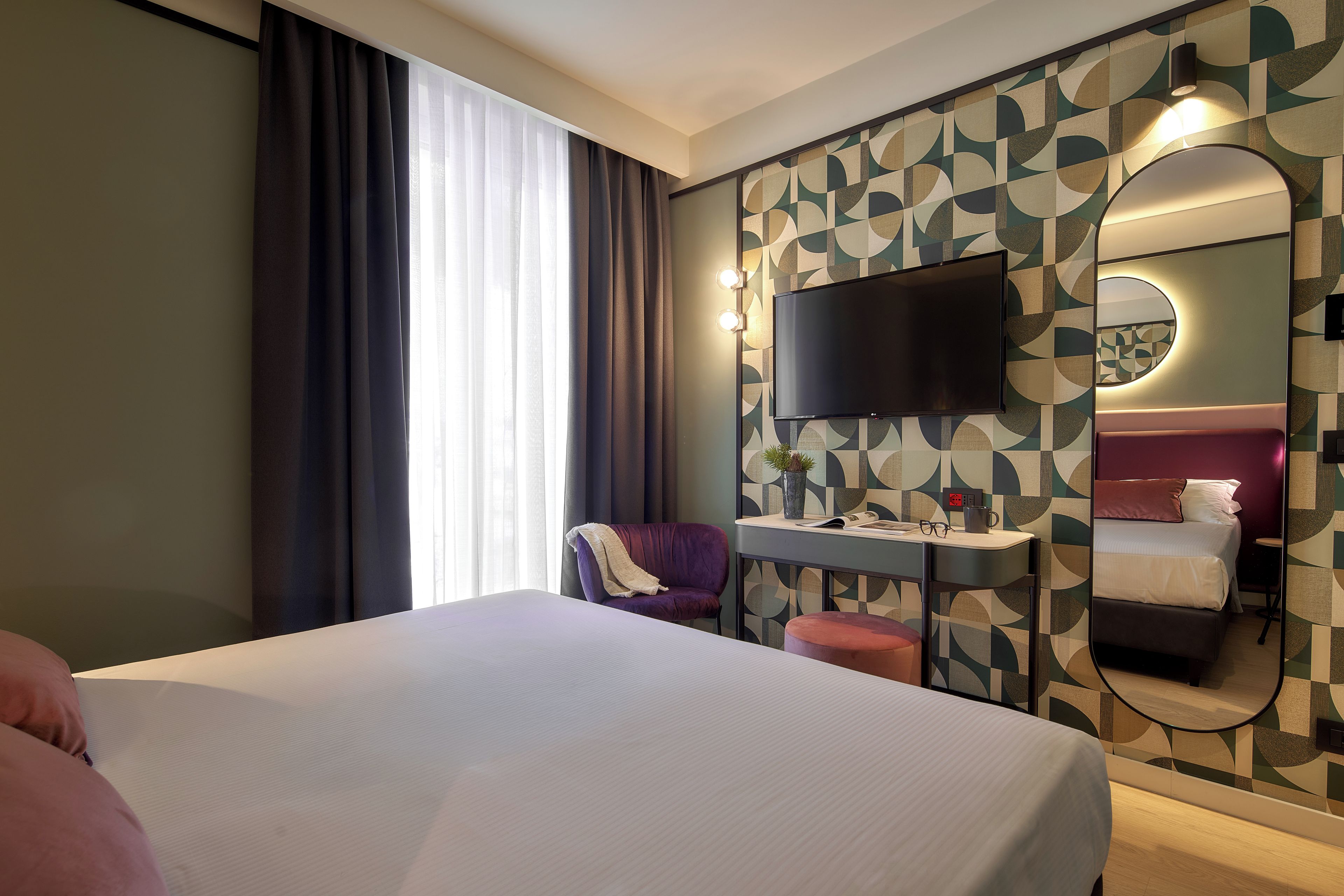A hotel room with a bed, a desk with a flat-screen TV, a chair, a mirror, and a patterned wall.