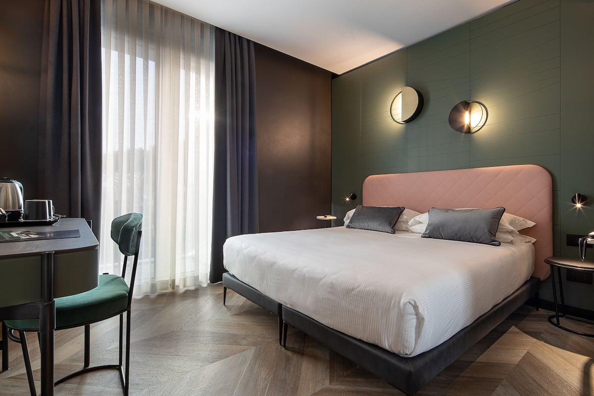 Habitación de hotel moderna con una cama grande, un escritorio con silla y vistas al exterior a través de cortinas traslúcidas.
