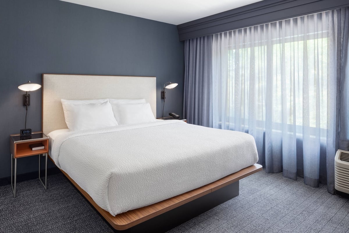 Una habitación de hotel moderna con una cama grande, lámparas de noche, una mesita de noche y un ventanal con cortinas translúcidas.