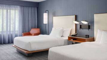 A hotel room with two beds, a desk with a telephone and a clock, a chair, and a wall-mounted lamp.