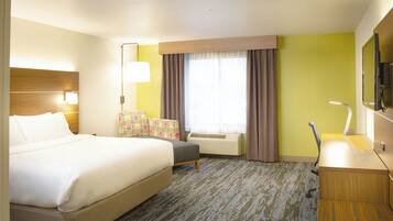 A hotel room with a bed, a desk, a chair, a window with curtains, an air conditioning unit, and a television.