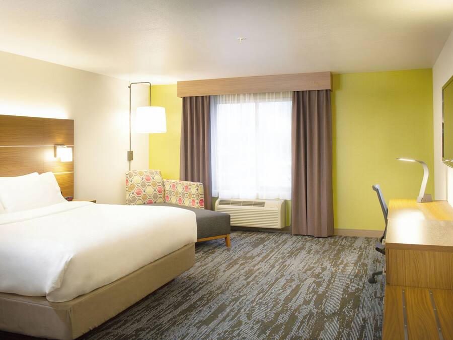 A hotel room with a bed, a desk, a chair, a window with curtains, an air conditioning unit, and a television.