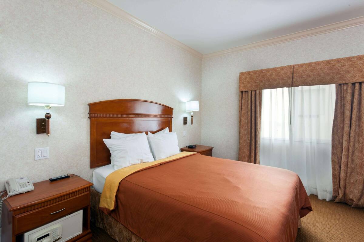 A hotel room with a wooden headboard, a bed with white linens, a brown throw blanket, a bedside table with a lamp, a phone, and a small drawer unit.