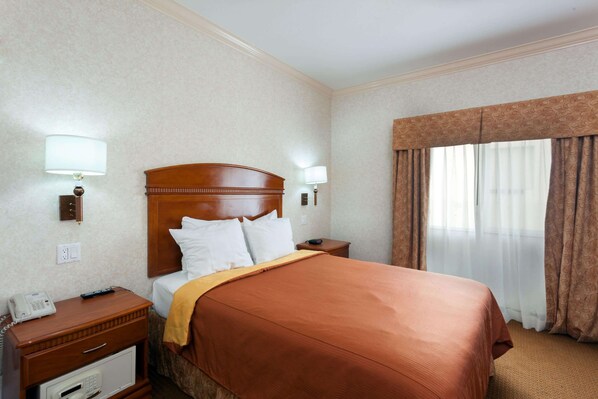 Habitación de hotel con cabecera de madera, cama con ropa blanca, manta marrón, mesita de noche con lámpara, teléfono y una pequeña cómoda.