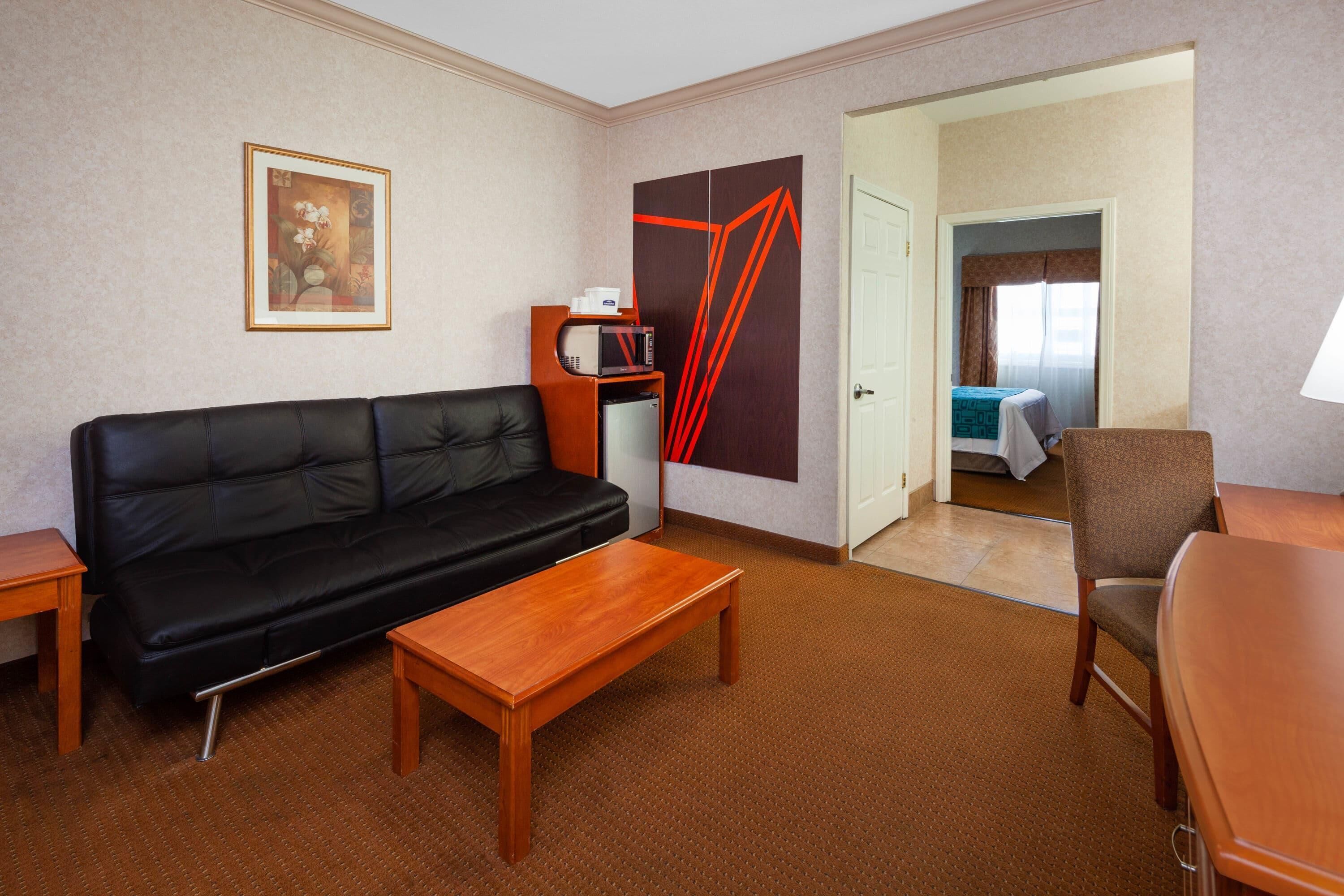 A hotel room with a black leather sofa, a wooden coffee table, a small side table, a microwave, a refrigerator, a painting, and a door leading to a bedroom with a bed and a chair.