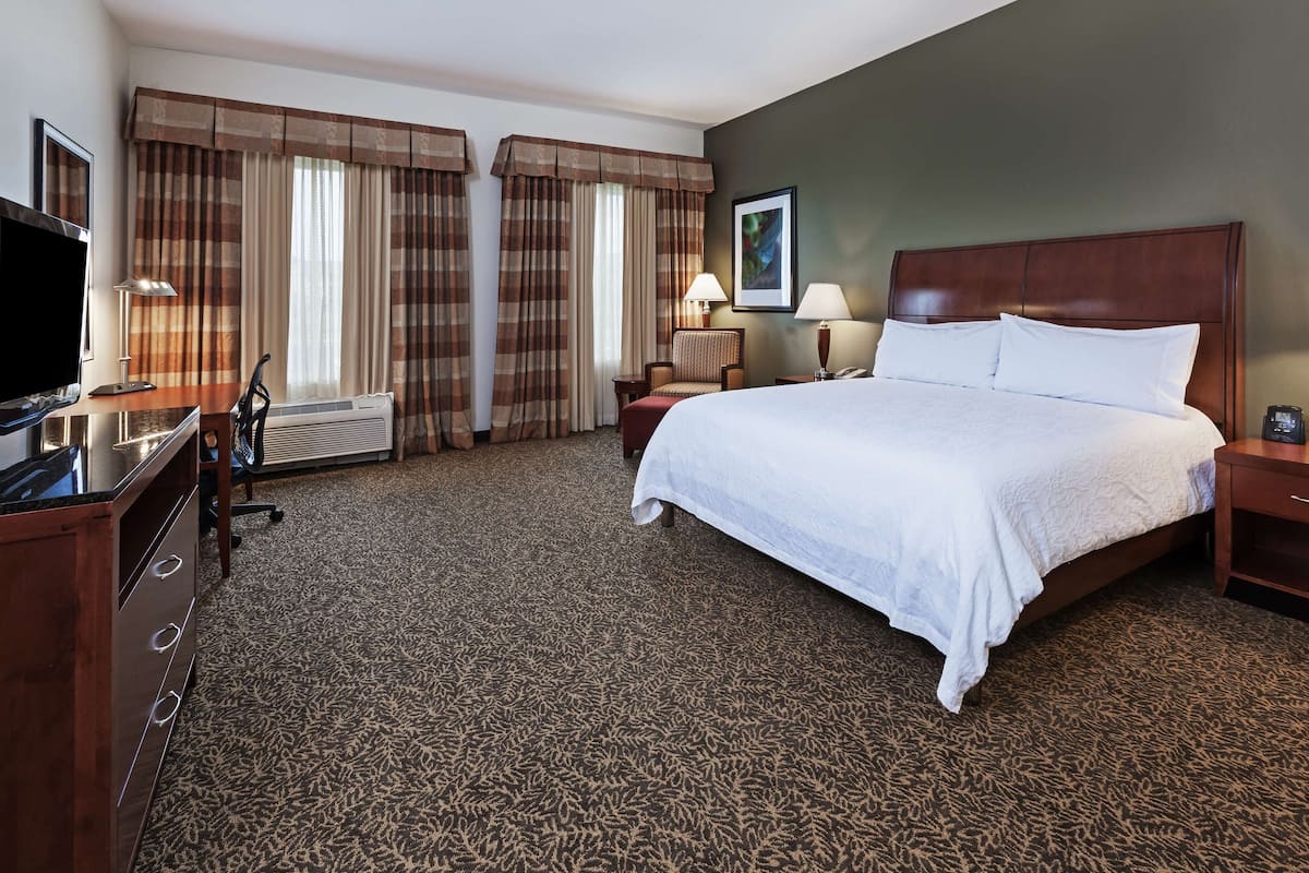 A hotel room with a large bed, a desk with a flat-screen TV, a chair, a lamp, and a framed picture on the wall.