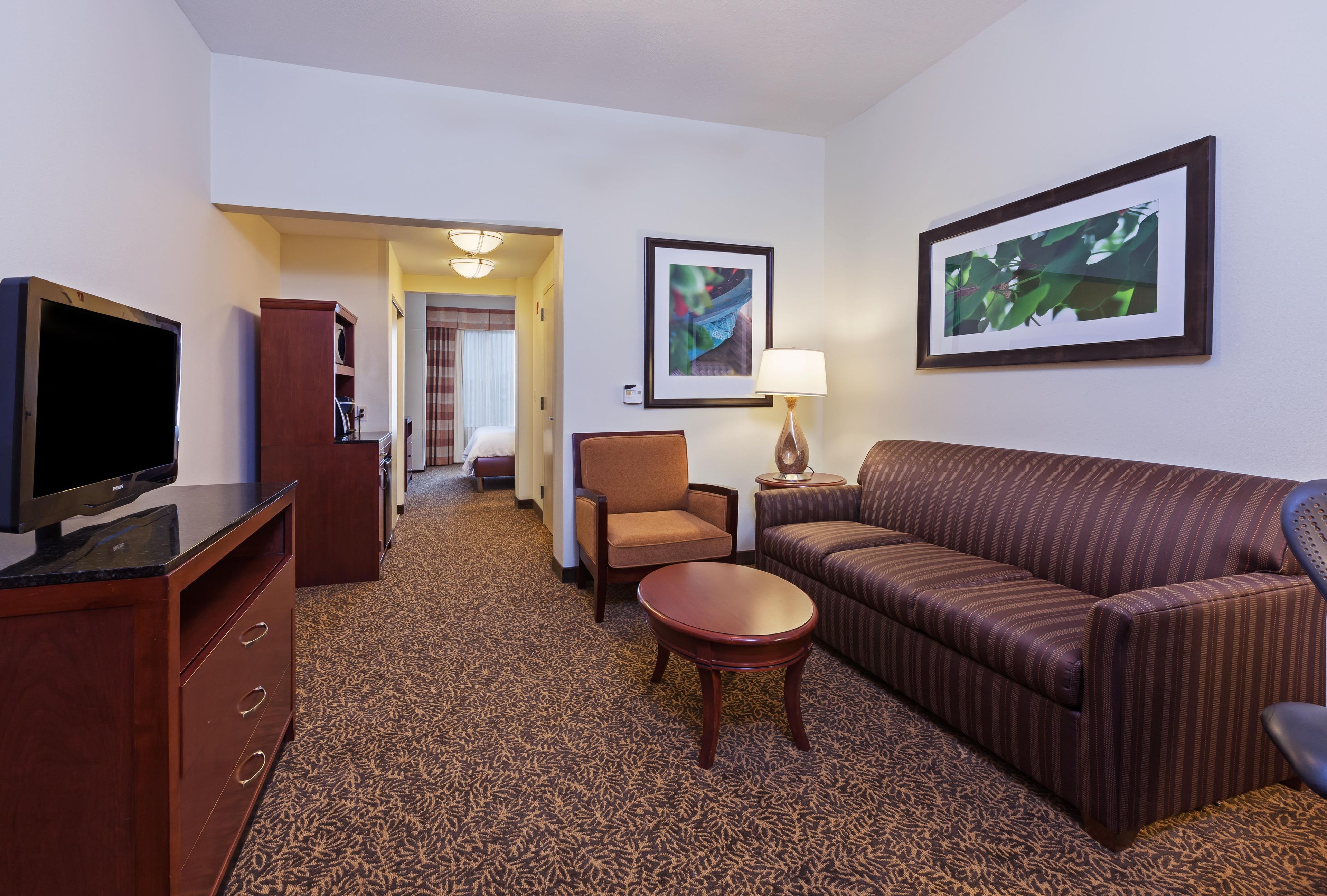 Quarto de hotel com televisão, sofá, poltrona, mesa pequena e cama ao fundo.