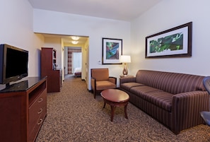 A hotel room with a television, a sofa, a chair, a small table, and a bed in the background. - Hilton Garden Inn Corpus Christi (Corpus Christi)
