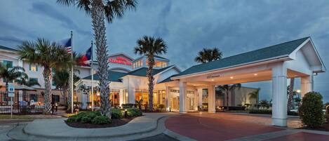 A hotel entrance with a covered walkway, palm trees, and flags.