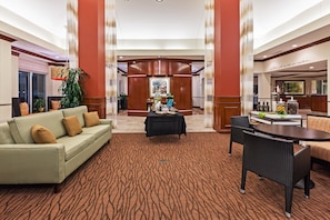 Un vestíbulo de hotel con una mesa de recepción, una zona de estar con sofá y sillones, y una mesa de comedor con estación de café.