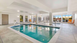 An indoor swimming pool with clear blue water, surrounded by white walls and a tiled floor. There are lounge chairs and a door visible in the background.