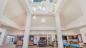 A spacious hotel lobby with a high ceiling, a central reception desk, and a dining area with tables and chairs.