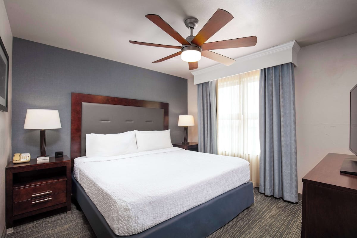 Una habitación de hotel con una cama grande, dos mesitas de noche con lámparas, un ventilador de techo y un televisor sobre un aparador.