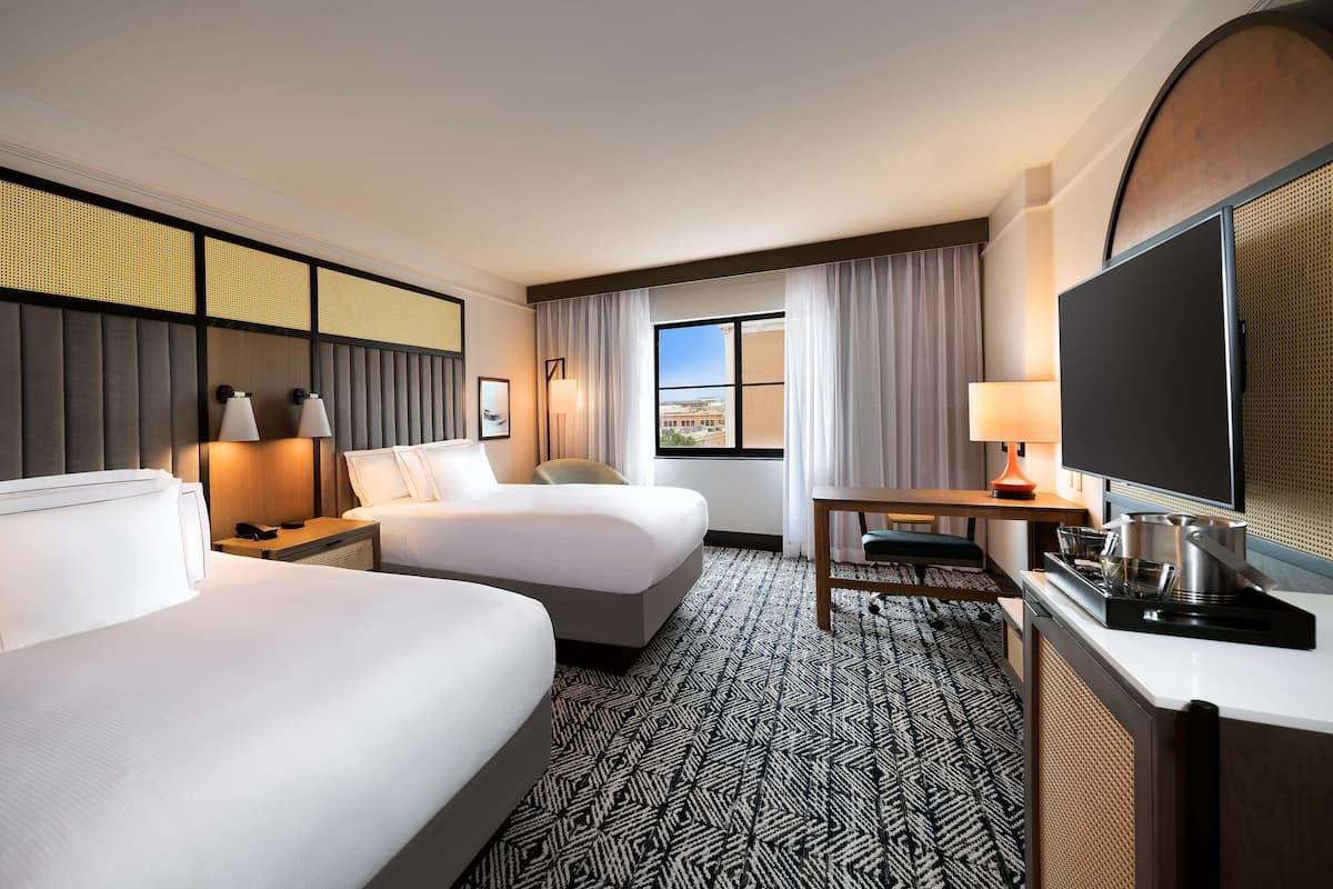 A hotel room with two beds, a desk, a television, and a window with curtains.