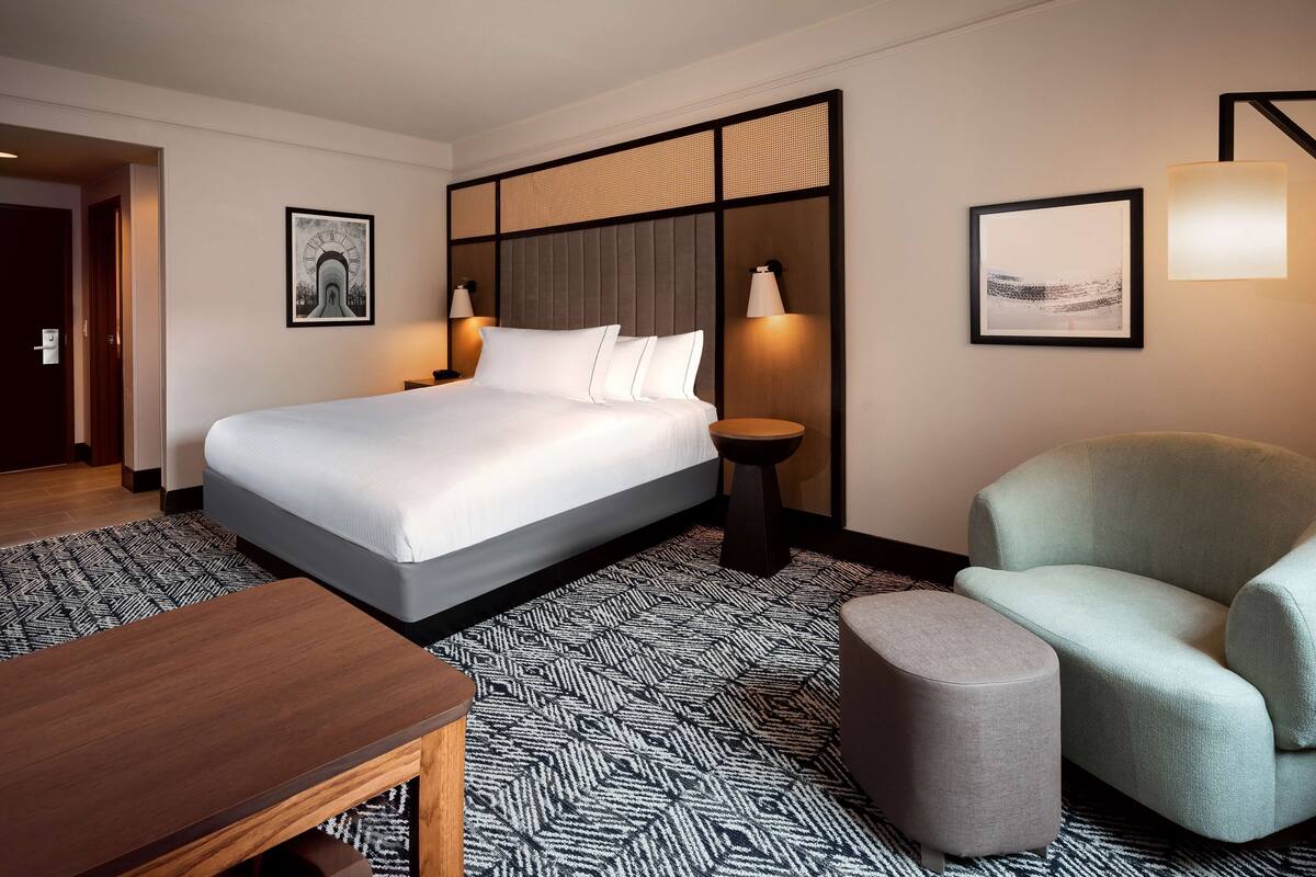 A hotel room with a large bed, a bedside table, a chair, and a small ottoman.
