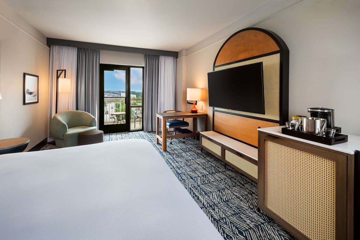 A hotel room with a large bed, a desk with a chair, a television, and a balcony with a view.