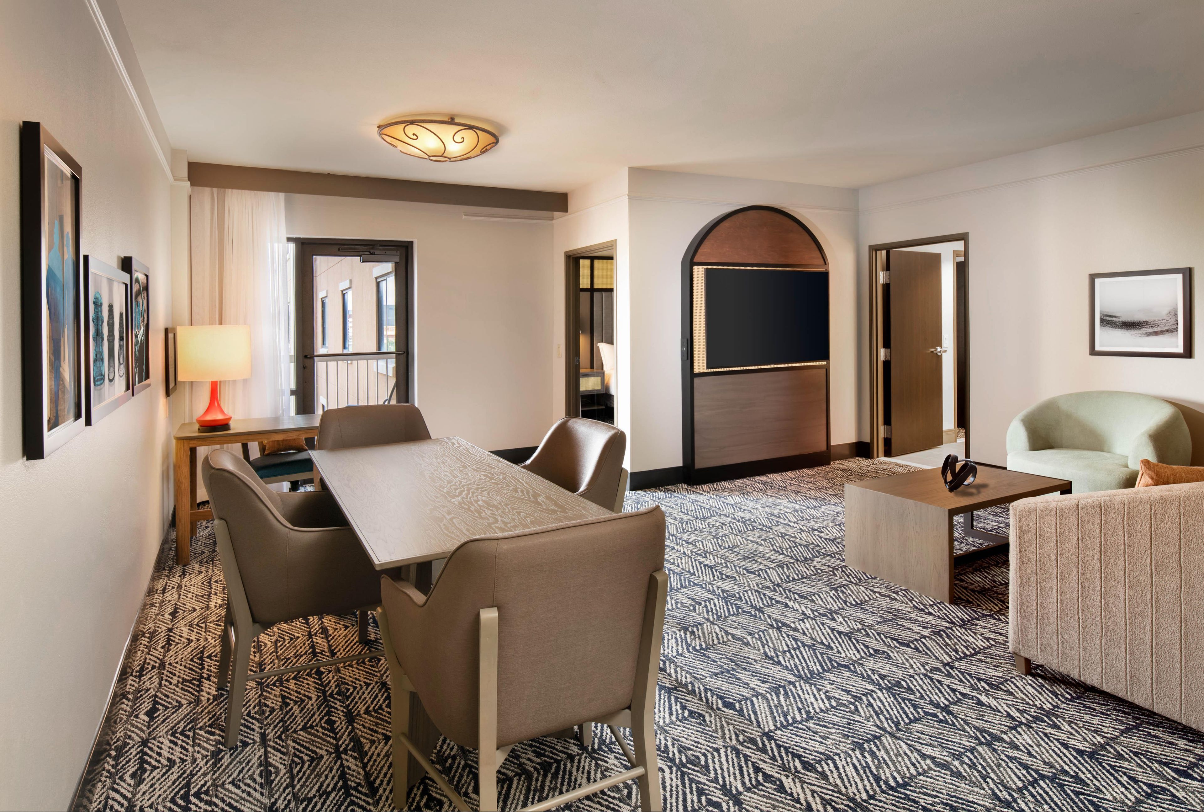 A hotel room with a dining table, chairs, a sofa, a TV, and a patterned carpet.