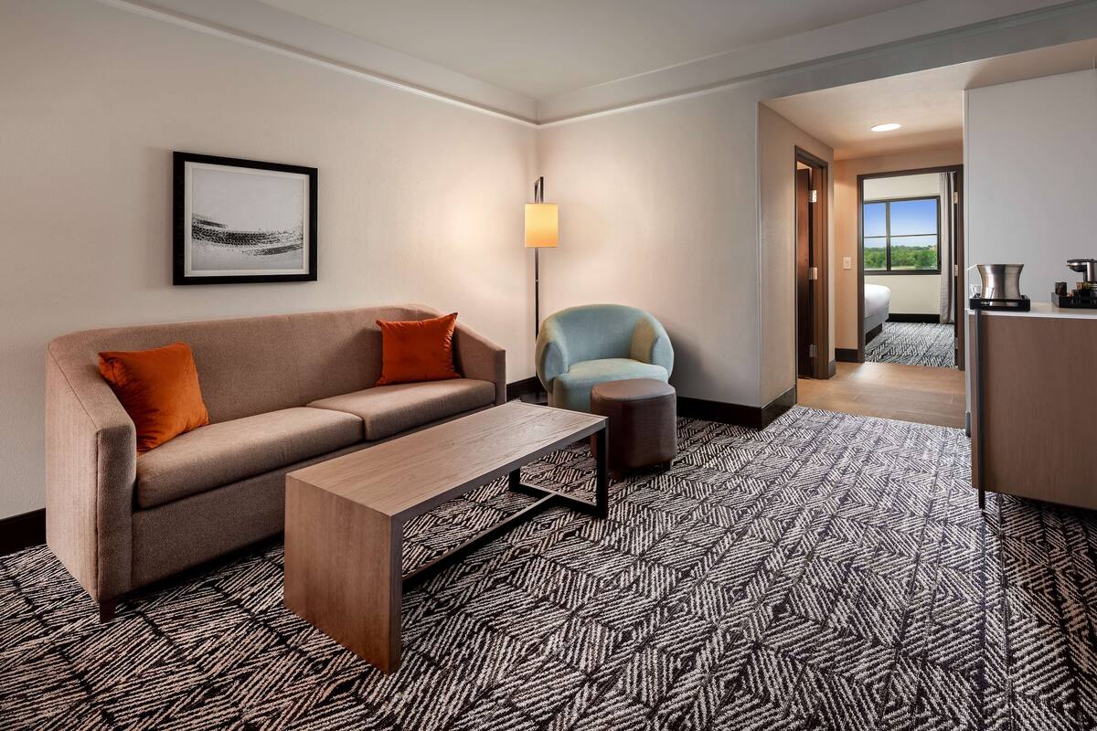 A hotel room with a sofa, a small table, a chair, a lamp, and a view of the outdoors.