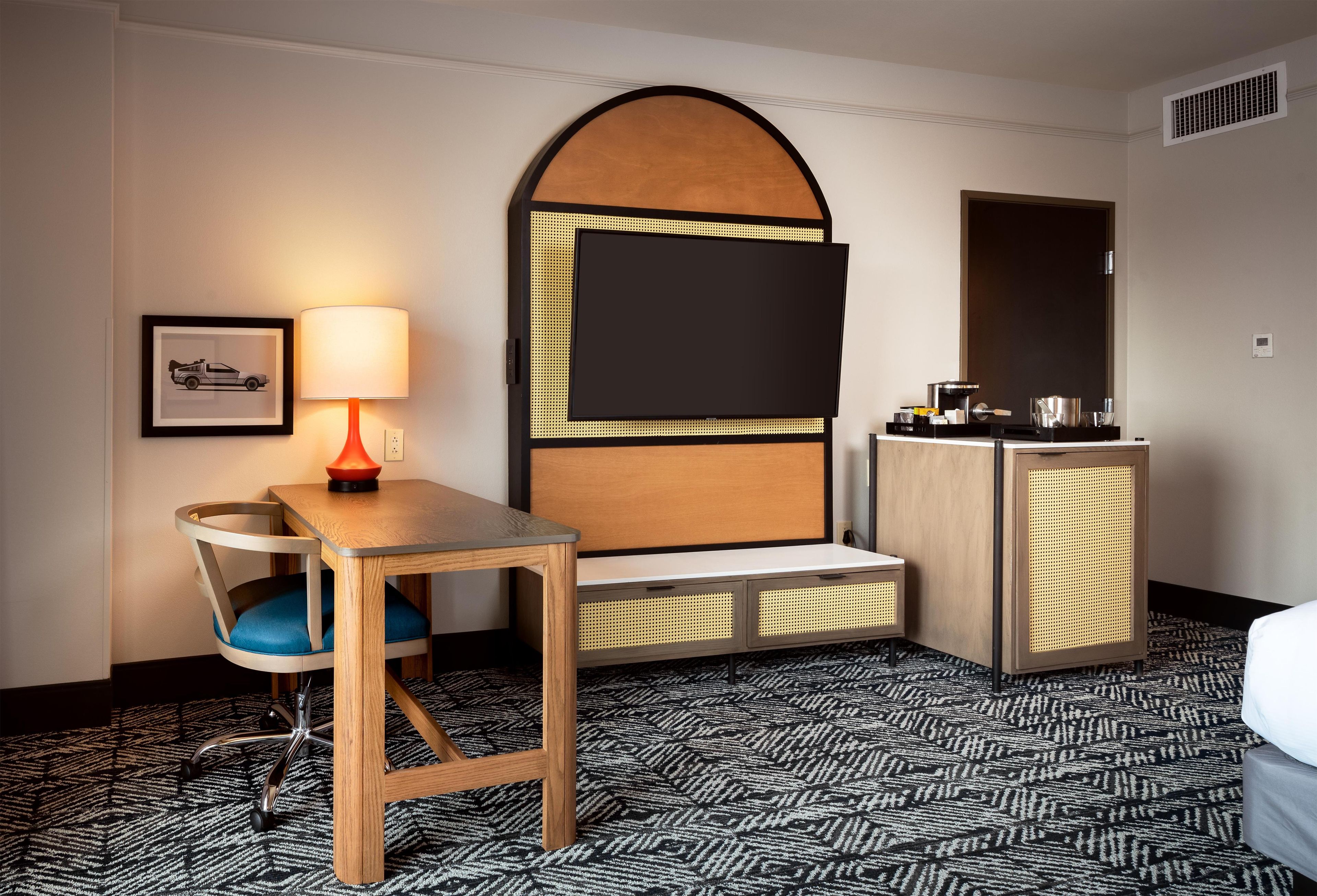 A hotel room with a wooden desk, a chair, a bed, a small cabinet, and a wall-mounted TV.