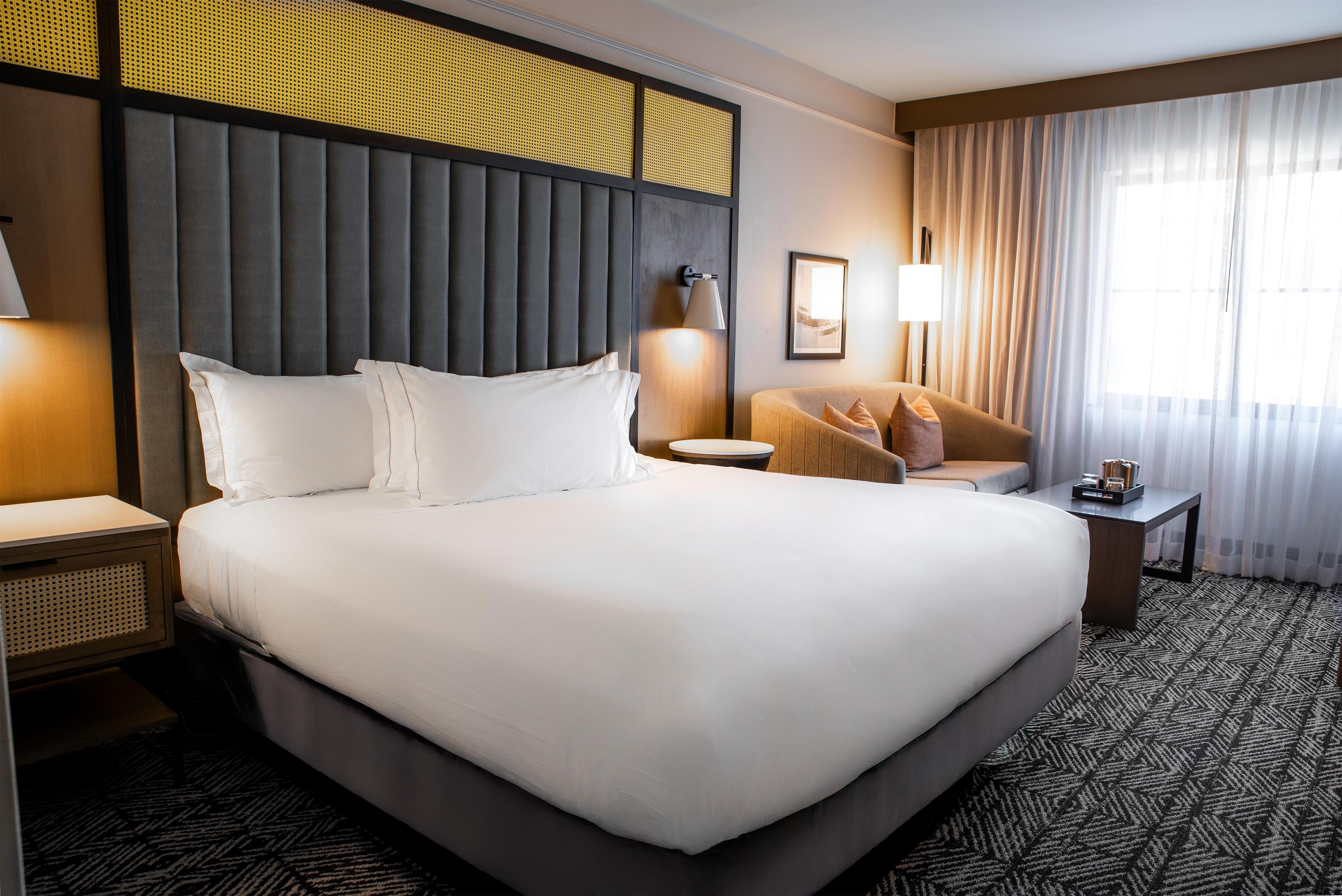 A hotel room with a large bed, a sitting area with a chair and a small table, a lamp, and a window with curtains.