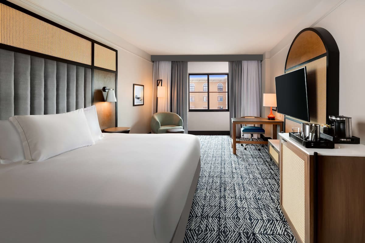 A hotel room with a large bed, a desk with a computer, a TV, and a view of buildings through the window.
