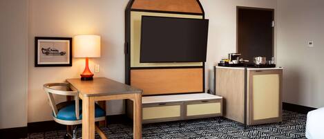 A hotel room with a bed, a desk with a chair, a television mounted on a wooden panel, a framed picture on the wall, and a patterned carpet.