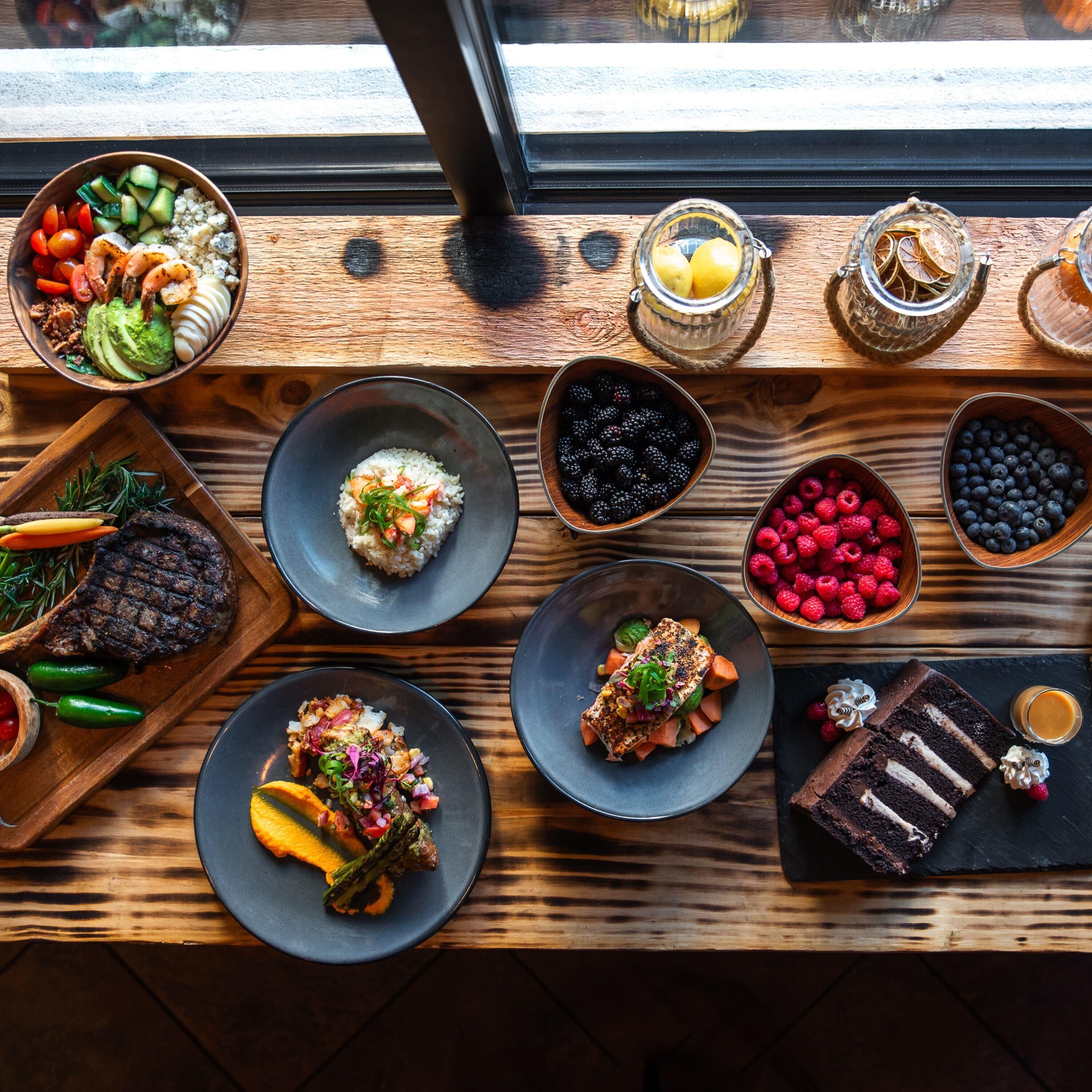 A variety of dishes on a wooden table, including grilled steak, salmon, and various fruits and vegetables.