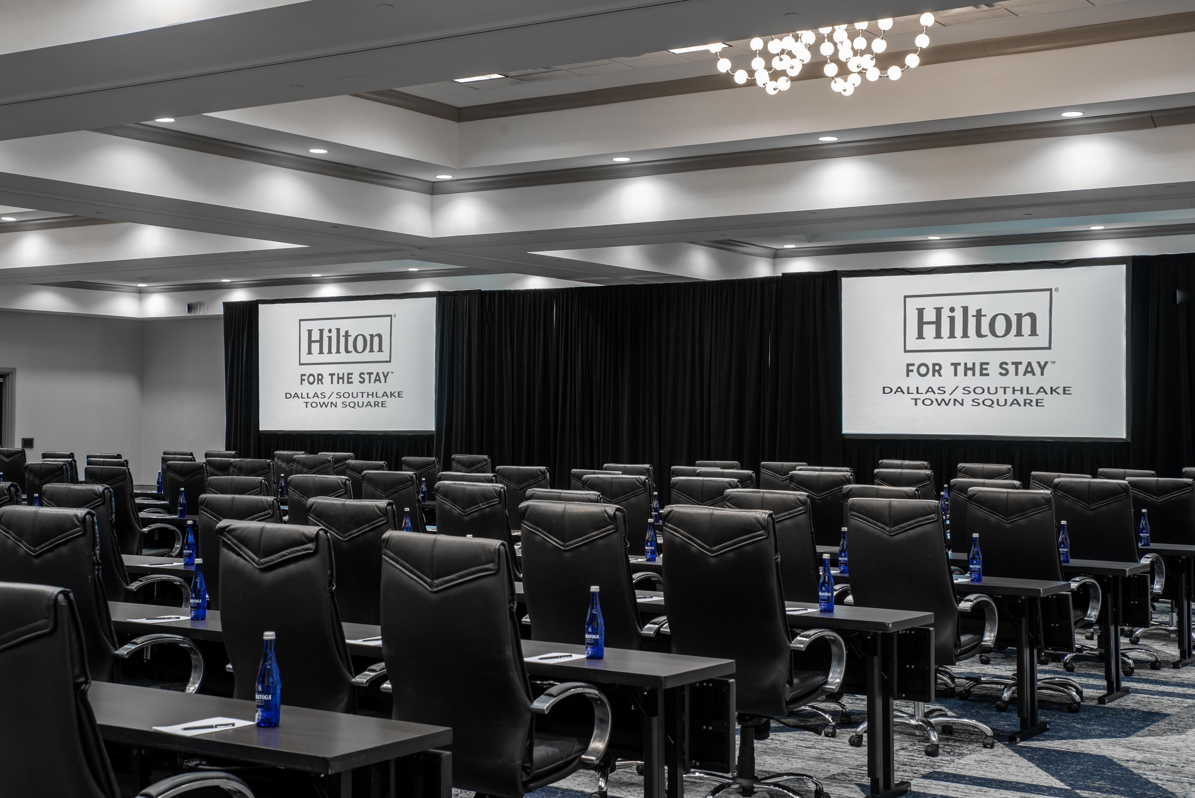 A conference room with black leather chairs, tables, and a large screen displaying the Hilton logo.