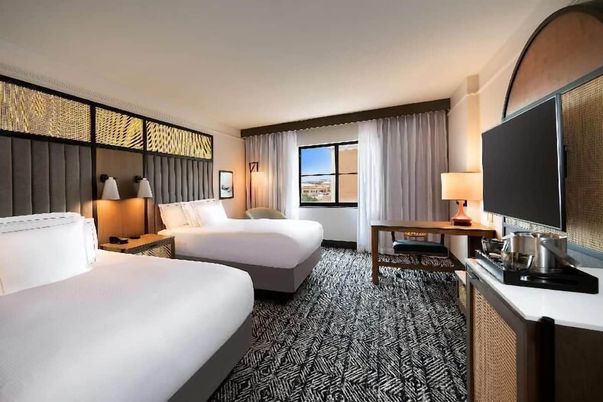 A hotel room with two beds, a desk with a TV, a window with curtains, and a patterned carpet.