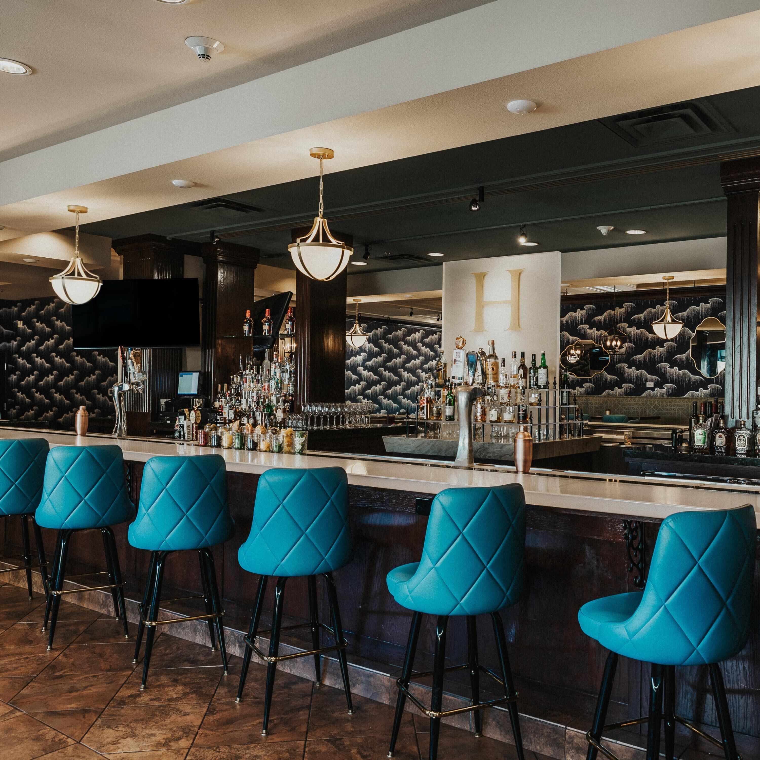 A bar area with multiple stools, a well-stocked bar counter, and a large H sign on the wall.