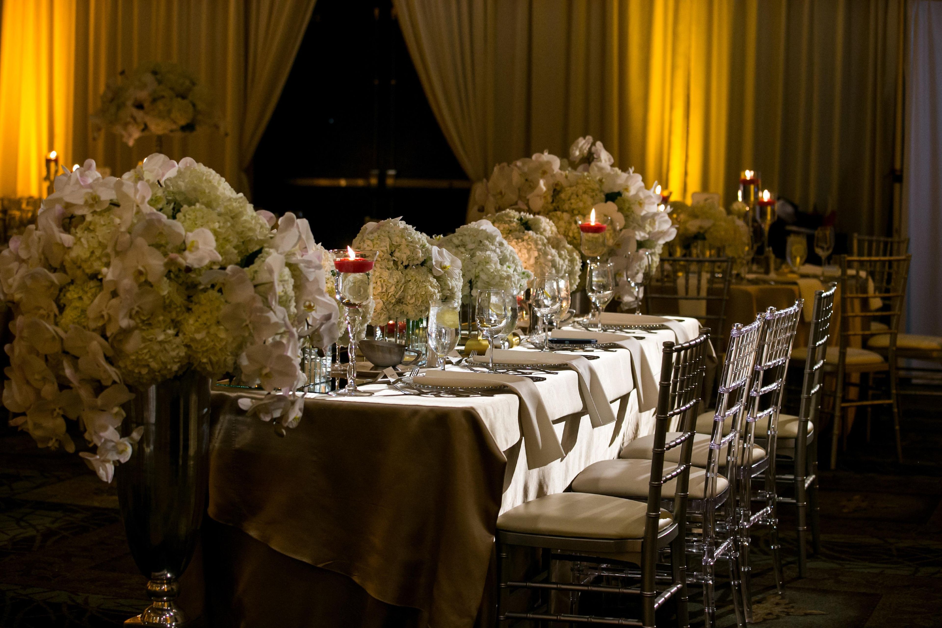 A formal dining setup with tables set for multiple guests, featuring floral centerpieces and candles.
