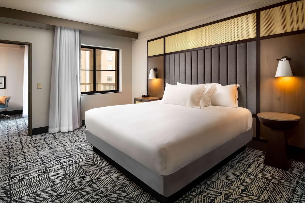 A hotel room with a large bed, two bedside tables with lamps, a window with a view, and a patterned carpet.