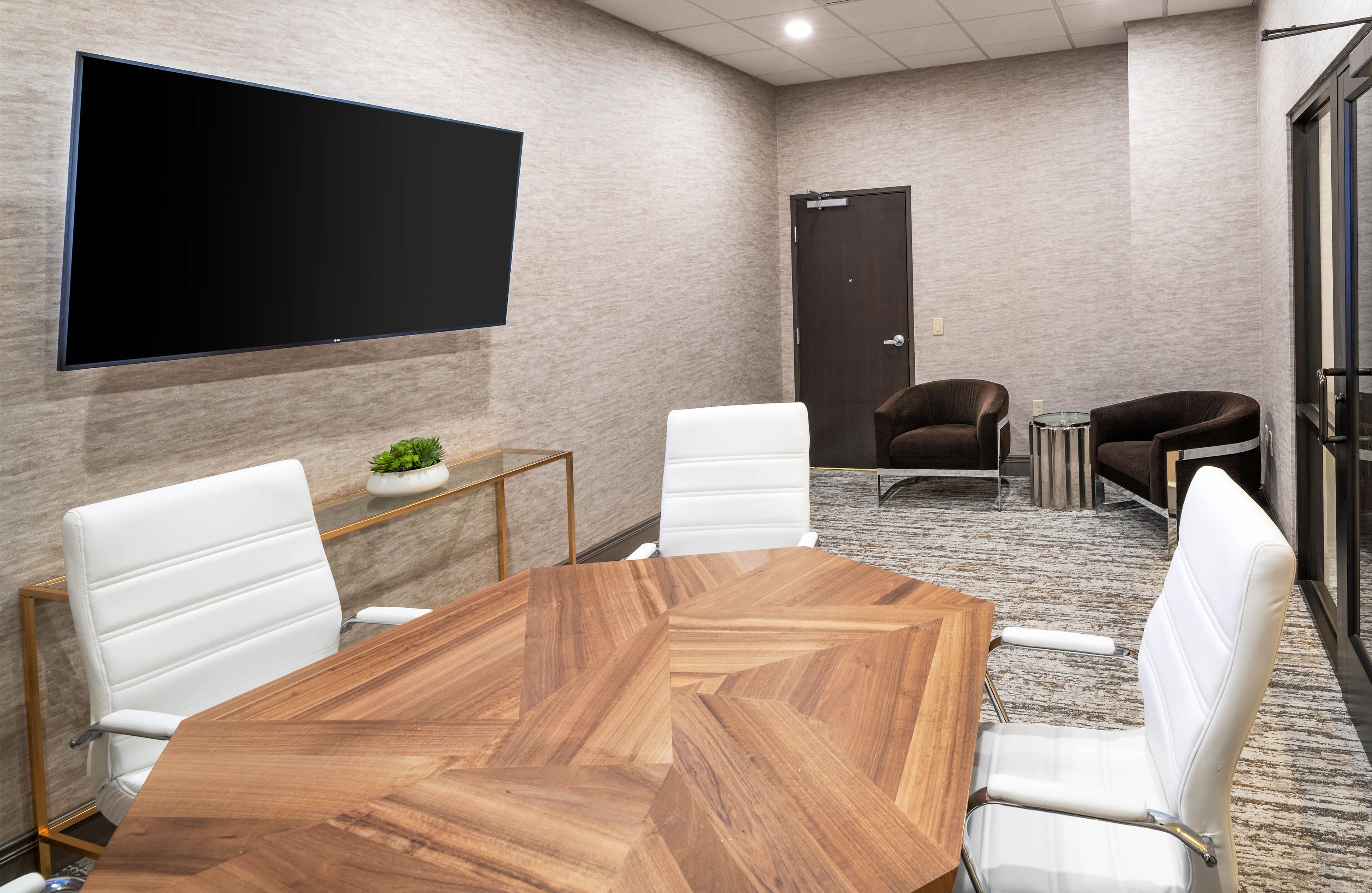 Une salle de conférence avec une grande table en bois, des fauteuils en cuir blanc, un téléviseur à écran plat fixé au mur et une porte à l’arrière-plan.