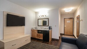 A compact hotel room with a flat-screen TV mounted on the wall, a small kitchenette with a microwave, a coffee maker, and a wooden cabinet. There is a dark-colored sofa and a small rug on the floor. - Holiday Inn Express Kalispell by IHG (Kalispell)