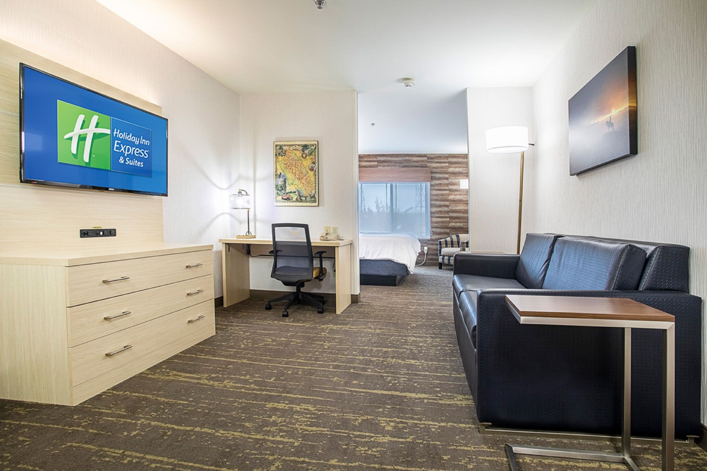A hotel room with a desk, a chair, a bed, a sofa, a TV, and a painting on the wall.