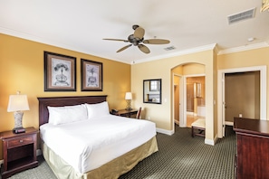 A hotel room with a large bed, two framed pictures on the wall, a ceiling fan, and a door leading to another room.