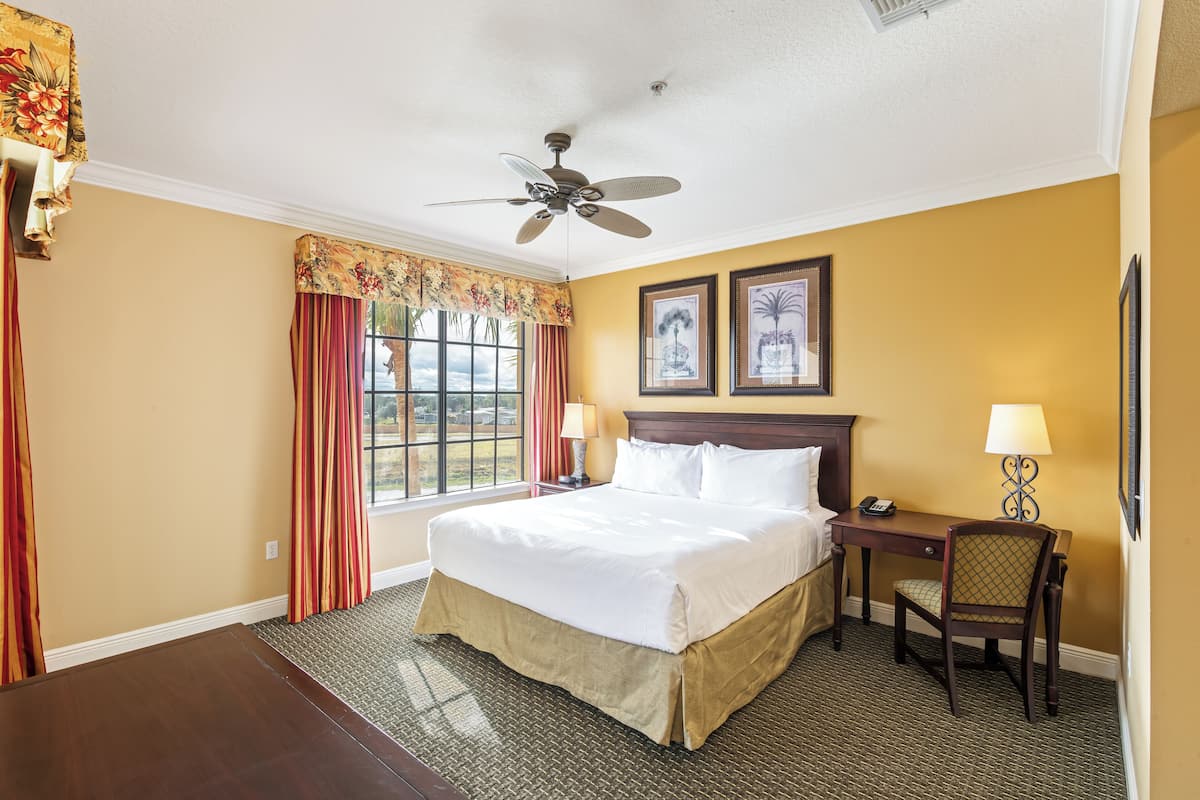 Ein Hotelzimmer mit Bett, Schreibtisch, Sessel, Deckenventilator und zwei gerahmten Bildern an der Wand.