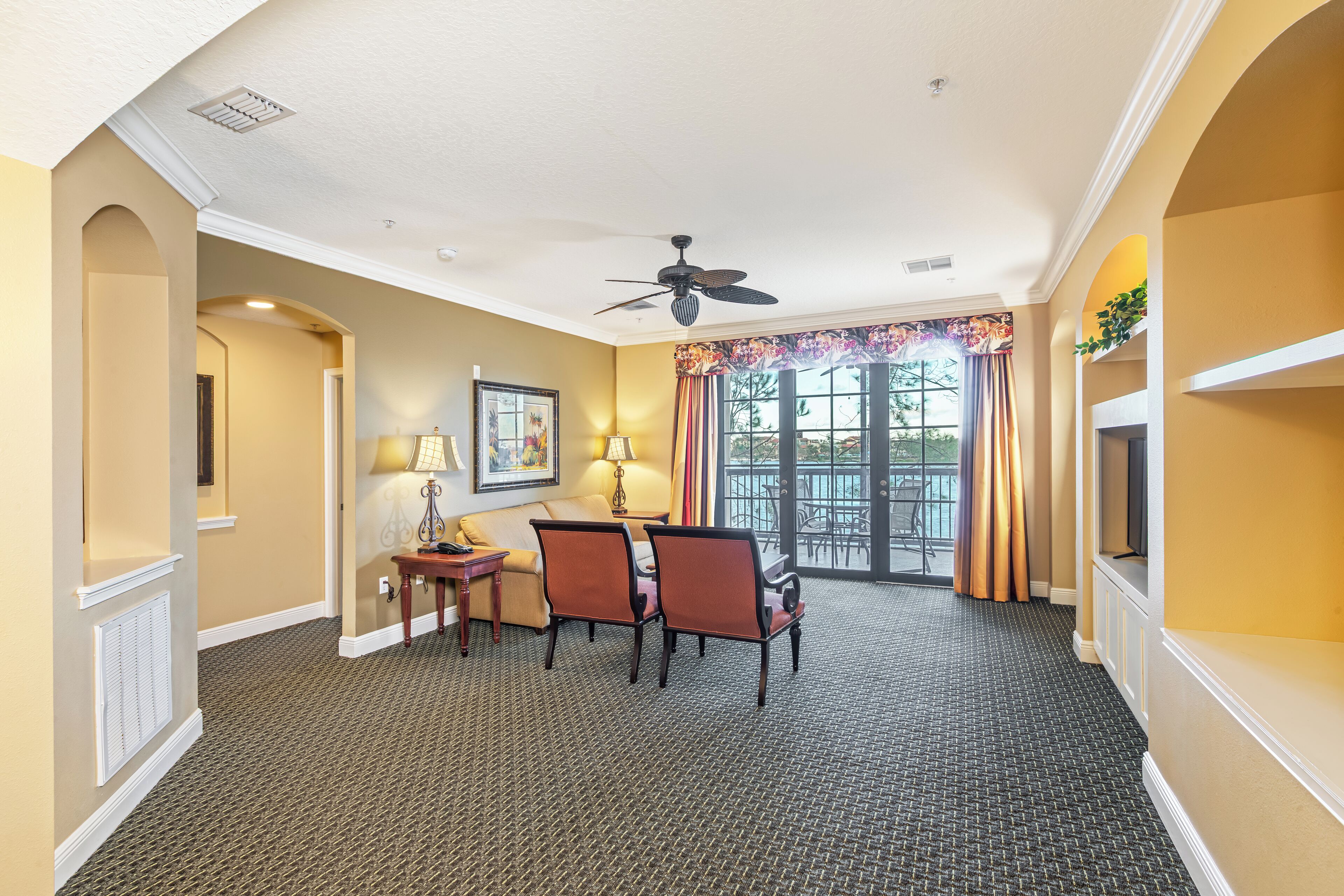 A hotel room with a dining area, a ceiling fan, a large window with curtains, and a view of the outdoors.