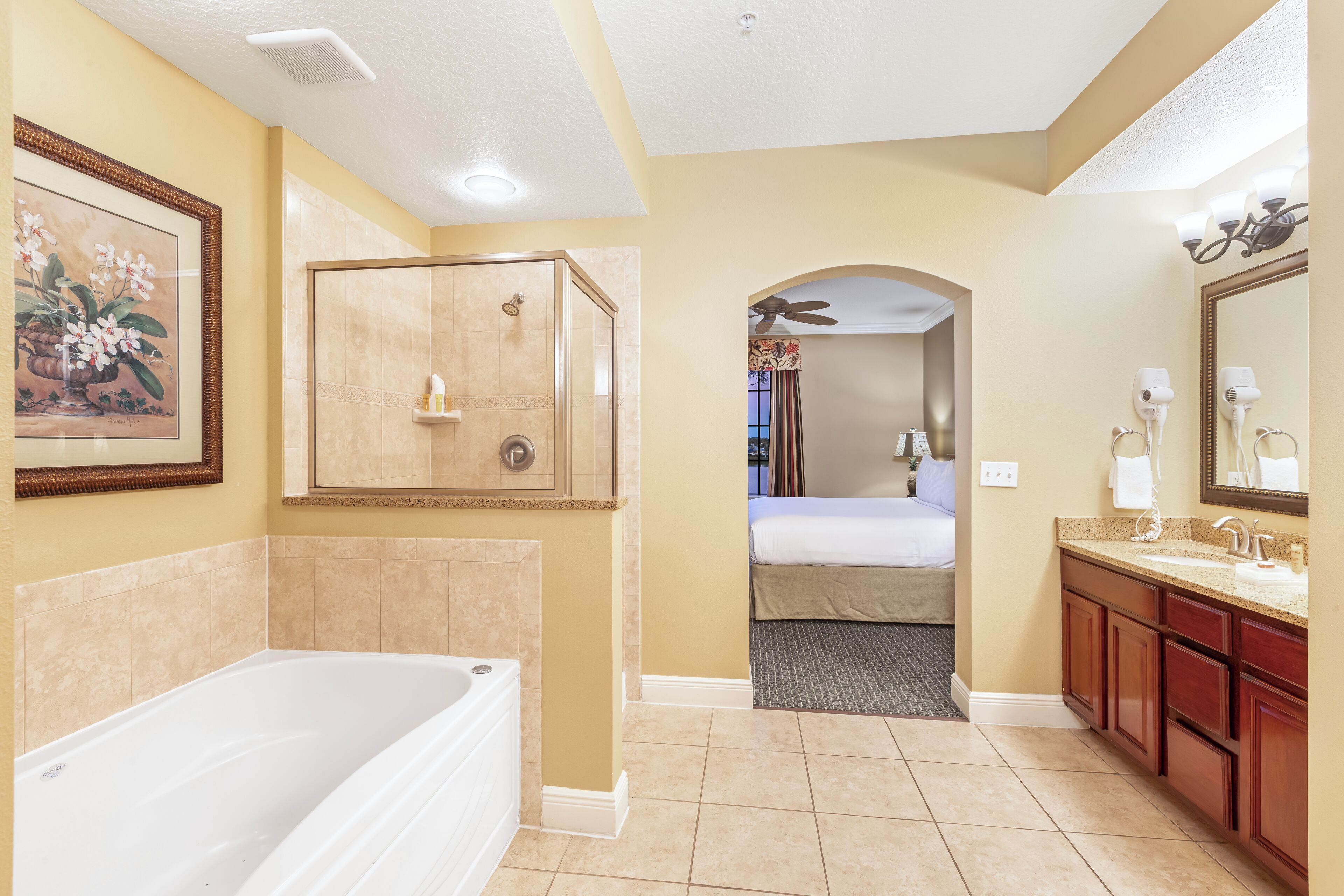 A bathroom with a bathtub, shower, vanity, and a bed in the adjacent room.