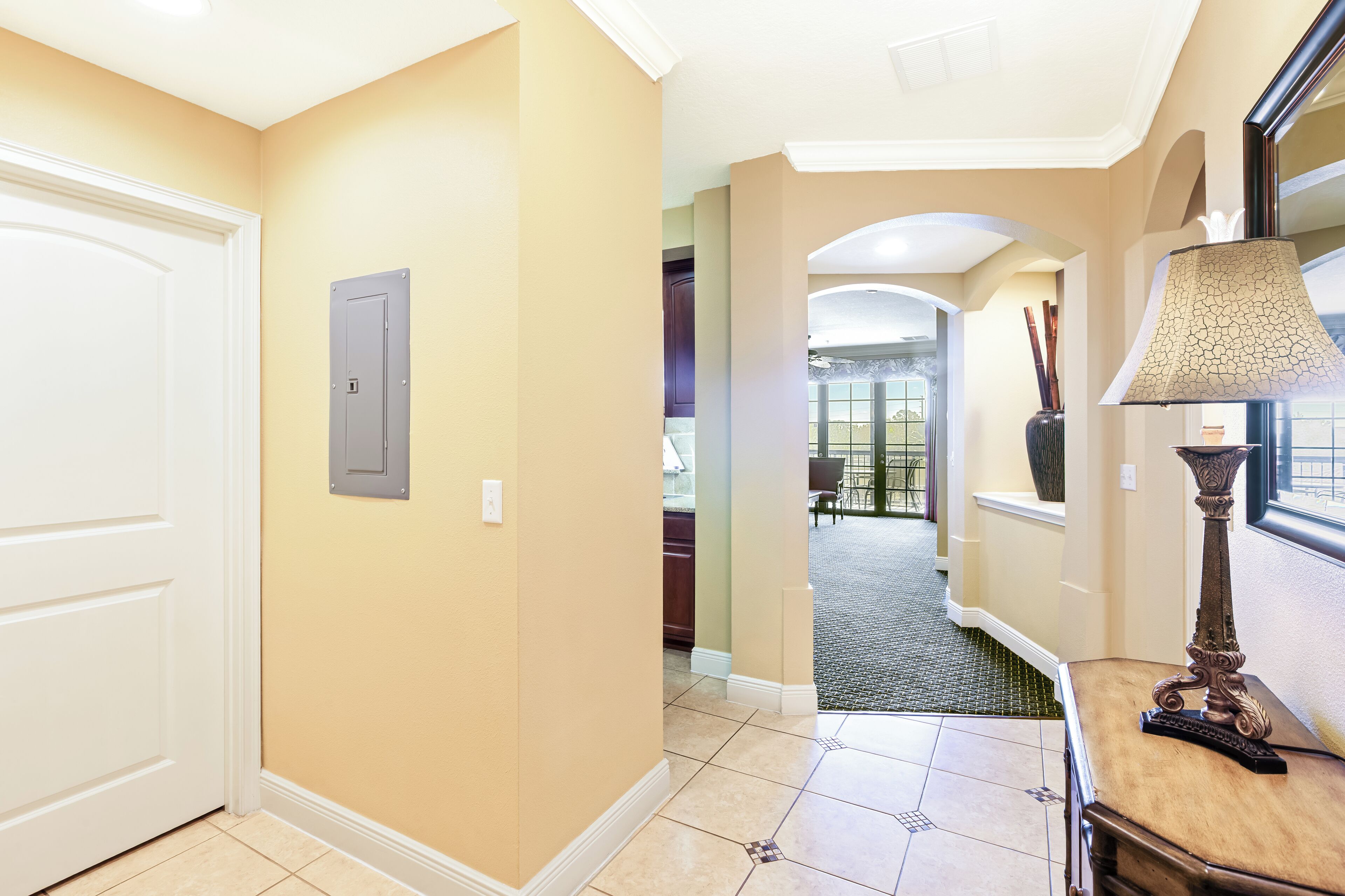 A hallway with beige walls, a white door, a grey electrical box, and a lamp on a wooden table.
