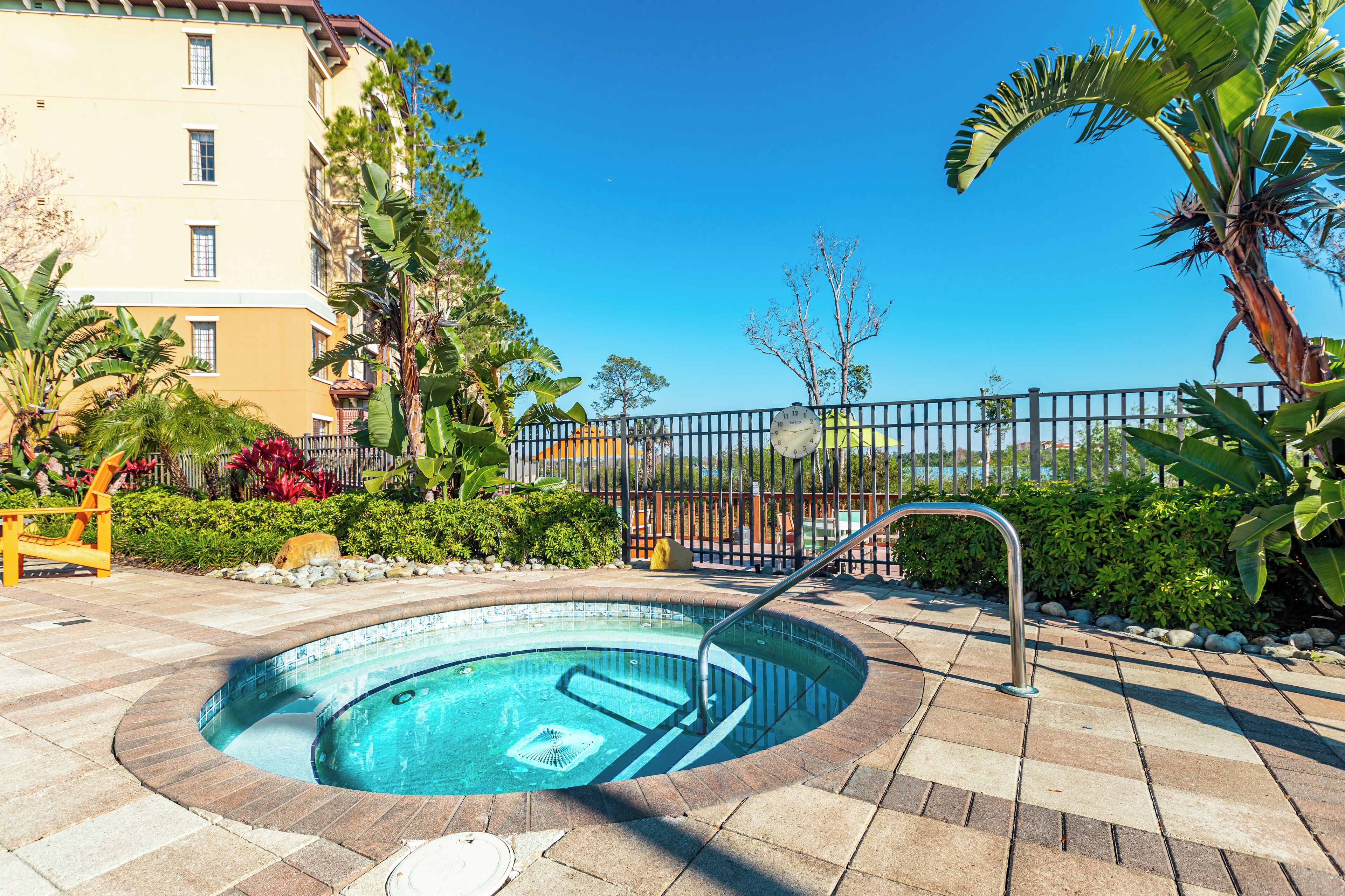 A circular hot tub with a metal ladder, surrounded by a paved patio with a fence and greenery.